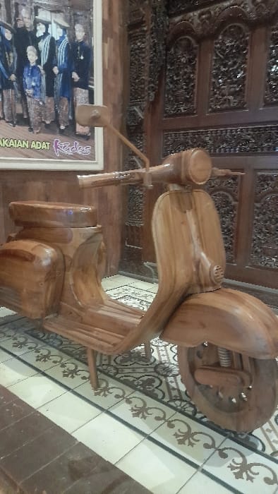 This image depicts an indoor scene in Glantengan, Indonesia. The central subject is a life-sized sculpture of a vintage Vespa-style scooter, meticulously crafted from light-toned, polished wood, complete with handlebars, seat, and wheels, standing on several small wooden support legs. The floor beneath is covered with ceramic tiles featuring a pattern of light-colored squares with dark outlines and interspersed darker tiles with elaborate floral or geometric designs. In the background, to the left of the wooden scooter, a large framed poster is visible, displaying a group of approximately eight individuals, including adults and a child, dressed in traditional Indonesian clothing. Partially legible text beneath the group reads "PAKAIAN ADAT Kedes." To the right, a dark wooden wall or partition with extensive and intricate traditional carvings forms the backdrop. No living individuals are present or engaged in any action within the frame.