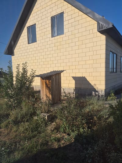 This image shows the exterior of a light yellow brick house under a clear blue sky. The house has a dark gray roof with visible gutters and trim. Two rectangular windows are visible on the upper part of the house's facade. On the side of the house, near the ground level, is a small, rustic wooden outbuilding with a corrugated roof. The outbuilding appears to be a shed or perhaps a toilet enclosure. In front of the house and around the outbuilding, there is overgrown vegetation, including bushes and tall grass, suggesting a rural or semi-rural setting. The foundation of the house is visible, made of concrete. Sunlight casts shadows on the side of the house and across the vegetation. The image does not show any people or evidence of recent activity. The location is specified as Bereke, Kazakhstan.