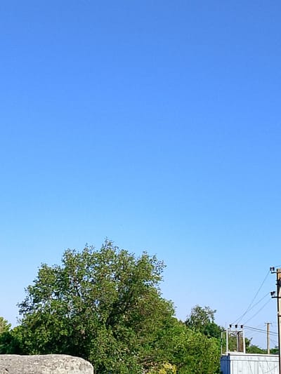 The image shows a clear blue sky above a line of trees. On the right side of the image, there is a utility pole with wires and a small electrical substation. In the foreground, a portion of a concrete structure is visible. The scene appears to be an outdoor setting in Pryvilne, Ukraine, possibly a rural or suburban area. There are no people or animals present in the image. The overall lighting suggests it is daytime.