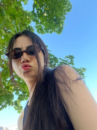 A young woman with long, dark hair and sunglasses puckers her lips for a selfie outdoors. She is positioned at a low angle, looking up towards the camera. Lush green tree leaves hang from above, partially obscuring the bright blue sky. The background suggests an urban or suburban setting, with a glimpse of rooftops and solar panels visible at the bottom left of the frame, consistent with the city of Dobřany, Czechia. The woman is wearing a dark top.