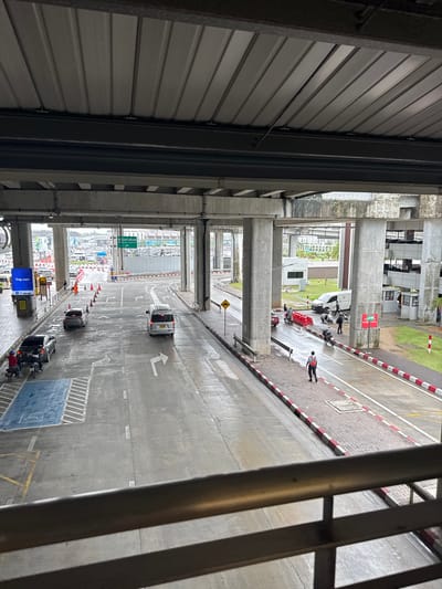 This image presents an elevated view of a bustling multi-level transportation hub, likely an airport drop-off or pick-up area, consistent with Tambon Sa Khu, Thailand. The scene is dominated by robust concrete support pillars and beams, forming an overhead structure. A corrugated metal wall runs along the left, while metal railings provide a partial frame on the right.
The ground level features a wet, paved road displaying various white markings, including directional arrows for turning left and proceeding straight. Adjacent to the road are pedestrian walkways with a distinctive brick-like pattern, delineated by red and white painted curbs.
Several vehicles are visible: a white van, a dark car, and multiple motorcycles or scooters, some with red luggage strapped on. Further down the road, another light-colored van or bus is seen. People are scattered throughout the scene; one individual in an orange safety vest stands on the pedestrian area, and others are near vehicles, suggesting activity related to arrivals or departures.
Notable details include a digital display screen showing "Trip.com" in the foreground. Several road signs are present: a yellow diamond-shaped caution sign with a left-pointing arrow, and green overhead directional signs with visible Thai script, likely indicating airport terminals or directions. Traffic cones are also placed along the road. The damp appearance of the road surface suggests recent rain or wet conditions, and the lighting indicates an overcast day.