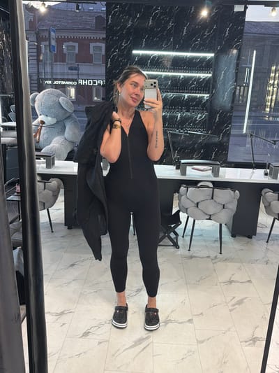 A woman stands in front of a mirror in what appears to be a nail salon. She is wearing a black, sleeveless jumpsuit with a zipper down the front, and black Crocs. She is holding a black jacket over her left shoulder. In the reflection of the mirror, a large gray teddy bear is visible to her left, and beyond that, a street scene with buildings. The floor is tiled with a marble-like pattern. The walls of the salon are decorated with black marble and shelves stocked with bottles, likely nail polish. The scene is lit by overhead lights and strip lighting on the walls. The woman is looking at her phone, which she is holding up to take a selfie.