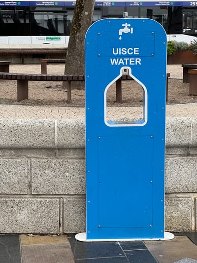 The image shows a blue outdoor water fountain located in Donegal, Ireland. The fountain is made of metal and has the word "UISCE WATER" written on it in white lettering, along with a graphic of a dripping faucet. The fountain is positioned on a paved area next to a stone wall. In the background, a bus is visible with various destinations displayed on its front, including "Donegal Town" and "Mountcharles". The environment appears to be outdoors, possibly a public space like a park or bus station, with benches and trees also present. The ground is wet, suggesting it may have recently rained.