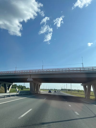 An eye-level, forward-facing shot captures a highway underpass on a bright, sunny day in Verkh-Tula, Russia. The sky is a clear, vivid blue, punctuated by scattered, fluffy white clouds. A large concrete overpass dominates the mid-ground, its gray structure supported by robust concrete pillars. A red railing runs along the top of the overpass, and streetlights are visible on either side. The highway below is paved and has white lane markings. Several cars are traveling on the highway, some are visible under the overpass and others further down the road. The surrounding landscape is characterized by green fields and trees, suggesting a rural or semi-rural setting. The overall impression is one of modern infrastructure amidst a natural environment.