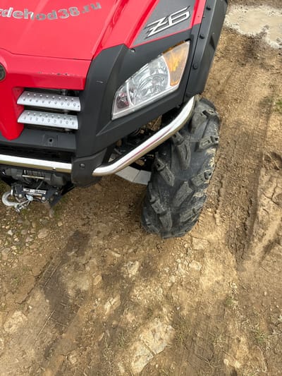 The image shows the front right side of a red all-terrain vehicle (ATV) parked on a dirt path. The ATV has a black fender, a silver bumper, and a large, knobby tire. The ATV also has a winch mounted on the front. The ground is covered in dirt, rocks, and tire tracks, indicating it is an outdoor, off-road environment. The sky is not visible in the image. The location is the city of Bolshaya Rechka, Russia.
