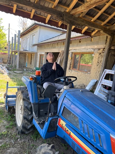 A person is sitting on a blue Iseki tractor, possibly a model 190, in an outdoor setting. The person is wearing a dark hooded sweatshirt and light-colored pants, with their hands together. The surrounding environment includes a dilapidated building with a wooden-framed window and a partially collapsed roof structure. Newer building materials are present, suggesting ongoing construction or renovation. The location appears to be a rural area, possibly in the vicinity of Rish, Bulgaria, based on the architectural style of the buildings. The ground is covered with grass and dirt.