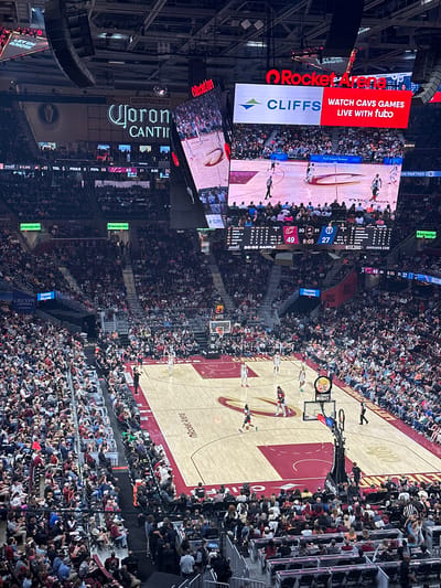The image captures a basketball game in progress at Rocket Mortgage FieldHouse, Cleveland, Ohio, United States. The arena is packed with spectators, mostly in casual attire. The basketball court is visible from an elevated angle, including players, referees, and team logos. Overhead, large digital screens display live footage, game statistics, and advertisements from sponsors such as Rocket Mortgage, Cliffs, Corona Cantina, Sherwin Williams, Huntington Bank, SeatGeek and Lecom. The score is displayed as Cavaliers 49 and the opposing team is at 27, with 8:05 remaining in the 2nd quarter.