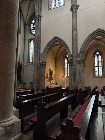 The image shows the interior of a large church with rows of dark wooden pews, some with red cushions, facing forward. Stained-glass windows in arched openings are visible, as are large stone columns and vaulted ceilings. In the background, a statue on an altar is partially visible. The scene is dimly lit, with light filtering through the stained glass. The image is a photograph of the interior of the Saint-Nicolas church in Ribeauvillé, France.