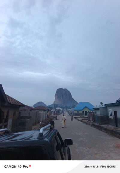 The image depicts a street scene in Wase, Nigeria. A large rock formation, likely Wase Rock, is visible in the background. The street is lined with buildings, some appearing to be residential homes. There are several people walking along the street. A vehicle is partially visible in the foreground, on the left side of the image. The sky is overcast. The camera settings are listed in the corner of the image as 23mm f/1.8 1/50s ISO893, taken with a Camon 40 Pro.
