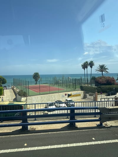 The image shows a coastal scene in Sitio De Calahonda, Spain, taken from a slightly elevated perspective, possibly from a bridge or a moving vehicle. The foreground features a blue metal railing, a road with white lines, and some debris. Beyond this, there is a concrete structure and a green bus. Further back, a tennis or paddle tennis court with red and green surfacing is visible, enclosed by a green fence. The sea is a prominent feature, with turquoise water meeting the horizon under a clear blue sky with a few clouds. Several palm trees are scattered around the court and the coastline. In the reflection on the glass, buildings and a logo are faintly visible. Two women are present near the tennis court area, one sitting on a ledge and another standing near a bus. Two white cars and one black car are parked in a lot near the tennis court.