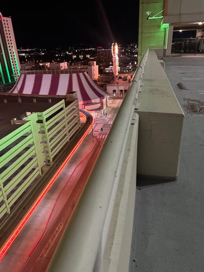 This nighttime, high-angle image captures a view of downtown Reno, Nevada, from an elevated structure, likely a parking garage, looking towards the Circus Circus Reno hotel-casino complex.

In the foreground, a concrete railing and barrier run diagonally from the lower right, partially obscuring the scene. The immediate concrete surface to the right of the barrier displays scattered debris and minor cracks. Below the railing, an illuminated street or elevated walkway extends into the midground, characterized by a continuous red LED light strip along its left edge. A single individual is visible walking or standing on this pathway near a white-marked crosswalk.

To the left of the pathway, a multi-story concrete parking garage is prominent, with its horizontal levels illuminated by a green light source. Adjacent and further left, the large, distinctive white and purple striped tent structure of the Circus Circus Reno is a central visual element. In the background, a tall building with vertical green lights stands on the far left, while the cityscape of Reno stretches into the distance under a dark sky, marked by numerous scattered artificial lights and silhouettes of other buildings, including one with a large, ornate illuminated sign to the right of the tent. The overall scene is static, with no overt actions or interactions observed beyond the solitary pedestrian.