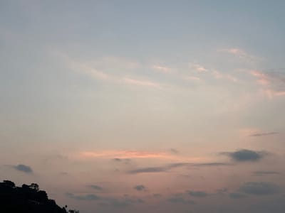 An outdoor photographic capture primarily features the sky during either dawn or dusk, constituting approximately 90% of the visual field. The upper sky exhibits a light blue-grey gradient, transitioning to soft pink, peach, and pale orange tones towards the lower frame. Cloud formations consist of scattered wisps and denser cumulus types; some appear translucent, while others in the lower and mid-sections are more opaque and darker grey, with patches of pink illumination. A dark, undifferentiated silhouette of a terrestrial landmass, consistent with a hill densely covered in vegetation, occupies the lower-left quadrant. No distinct flora or fauna details are discernible within this silhouette. A single, small, indistinct bright point is faintly visible along the lower edge of the landmass, its nature undetermined. No human subjects, specific man-made objects other than the aforementioned point, or any dynamic actions or interactions are present within the frame. The geographical context for this scene is the city of Preah Sihanouk, Cambodia.