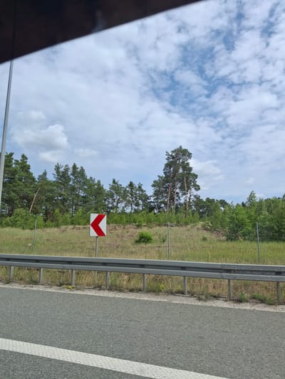 The image displays a roadside scene with a curve warning sign. The sign, a white rectangle with a red arrow pointing left, is positioned to the right of a metal guardrail. Beyond the guardrail, a grassy embankment is topped with a dense line of evergreen trees. The sky above is partly cloudy with scattered white clouds against a blue background. The foreground shows an asphalt road with a white line marking its edge. The location is specified as Brzoza, Poland.