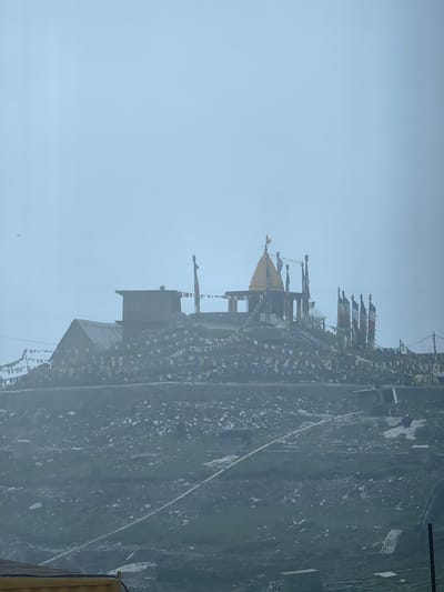A view of a religious site on a hillside in Marhi, India. The site features a temple with a golden spire and several flags, surrounded by prayer flags strung across the hill. The landscape is rocky and covered with sparse vegetation, with a pale, overcast sky dominating the upper portion of the image. In the foreground, there is a section of a wooden structure.