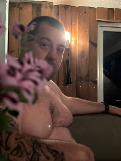 The image depicts a man in a room with wooden walls. He is shirtless and has tattoos on his arms and chest. He is holding pink flowers in the foreground, obscuring part of his face. A black flashlight is hanging on the wall behind him. The room appears to be indoors, possibly a cabin or a basement. The man is looking towards the camera. The image does not provide enough information to determine the specific location in Heinebach, Germany.
