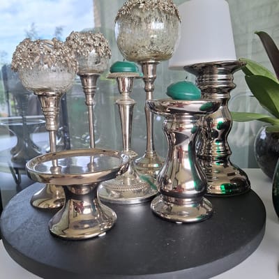 The image shows a collection of ornate silver candle holders and green candles arranged on a dark, round surface. In the background, a white lampshade is visible. The candle holders vary in design, with some featuring decorative floral elements and others having ribbed bases. The overall aesthetic suggests an indoor setting, possibly a home or a decorative display. There are no people visible in the image. The context of Mechelen, Belgium is not evident from the visual information in the image.