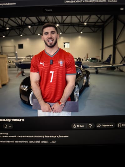 The image shows a screenshot of a YouTube video. In the foreground, a man is standing and looking at the camera. He is wearing a red soccer jersey with the number 7 on it, white shorts, and a red watch. He is holding a dark gray rectangular object in his hands. Behind him, there is a black sports car and several small airplanes in what appears to be a hangar or an aircraft hangar. The background is a bit blurred, but the overall setting seems to be an indoor space with industrial elements like metal beams and lights on the ceiling. The YouTube interface elements are visible at the top and bottom of the screenshot, including the video title "ТАМАЕВ КУПИЛ У РОНАЛДУ BUGATTI - YouTube" and playback controls. The text below the video shows Russian language labels like "Поиск" (Search), "53 тыс." (53 thousand), "Поделиться" (Share), and other video description text. There is no visual information in the image to suggest the location is the city of Демихово, Russia.