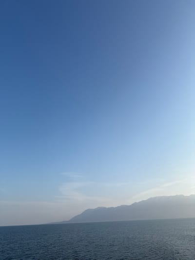 The image presents a panoramic view of a coastal environment in Kato Karioutes, Greece. The scene is divided into a clear blue sky occupying the upper two-thirds and a body of water in the lower third. The sky exhibits a gradient from a deeper blue at the top to a lighter, somewhat hazy blue near the horizon, particularly on the right, with sparse, thin white cloud formations visible in the mid-left to central horizon area. The water, likely a sea or large bay, is dark blue-green and features subtle ripples across its surface, indicating calm conditions. A lengthy, hazy grayish-blue mountain range or distant coastline stretches horizontally across the middle of the frame, emerging from the water. Patches of white, possibly snow or light-colored rock, are discernible on some of the higher peaks of the mountain range, predominantly towards the right side. No individuals, man-made objects, or active interactions are visible.