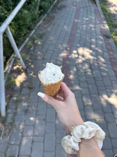 A person is holding a partially eaten ice cream cone in their right hand in Mamaivtsi, Ukraine. The ice cream appears to be vanilla with dark chocolate chips. The cone is a waffle cone. The person's hand has long, well-maintained fingernails painted white. A scrunchie made of a light-colored, shiny fabric is worn around their wrist. The background shows a paved walkway made of gray and red rectangular stones, with dappled sunlight filtering through trees. A metal railing and green foliage are visible on the left side of the image.