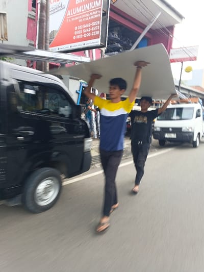 Two men carry a large white sheet above their heads while walking on a street alongside vehicles. The man in the foreground wears a blue and yellow shirt with dark pants and sandals. The second man wears a dark shirt and pants with sandals. A black van is partially visible on the left. In the background is a white van and a sign that reads "BINTANG GONYA ALUMUNIUM" with a Bandung address and phone numbers. The scene appears to be in an urban area.