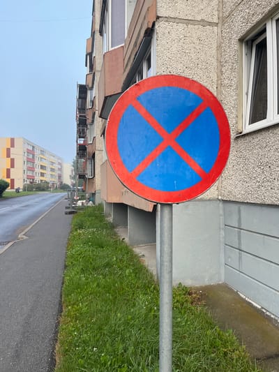 A no parking sign is situated on the side of a street in Maardu, Estonia, next to a grey concrete building. The sign, a red circle with a red "X" on a blue background, is mounted on a grey metal pole. The street is asphalt and appears wet, with a strip of green grass bordering the sidewalk. In the background, several apartment buildings with colorful facades are visible under a cloudy sky. Construction scaffolding is attached to the side of the building near the sign. There are no people or vehicles in the immediate vicinity.