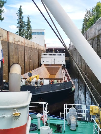 The image depicts a lock chamber in the city of Belousovo, Russia, where two barges are positioned. The foreground shows the deck of a green vessel, partially obscured by a large white cylindrical object. A white railing with vertical supports runs along the edge of the deck. Further into the chamber, a dark-colored barge with Cyrillic lettering reading "ПЕВСКИН-13" is visible, featuring a large, angled, rust-colored cargo hold. Behind this barge, a larger white vessel with multiple windows is docked. The lock chamber walls are constructed from large, textured concrete blocks, with water reflecting the sky and vessels. In the background, trees and a modern building with blue window frames are present under a cloudy sky. The scene suggests the ongoing operation of a lock for maritime transport.