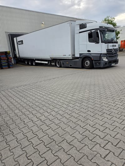 A white semi-trailer truck with the logo "Select Cargo Service" is parked at a loading dock of a large, corrugated metal building. The truck is partially backed into the building's entrance. The truck's cab is white with a black grille and a Mercedes-Benz logo. A license plate reading "GG K 5405" is visible on the front of the truck. To the left of the truck, a stack of colorful pallets is visible. The ground is paved with interlocking gray concrete blocks. The sky is overcast. This scene is located in Zeppelinheim, Germany.