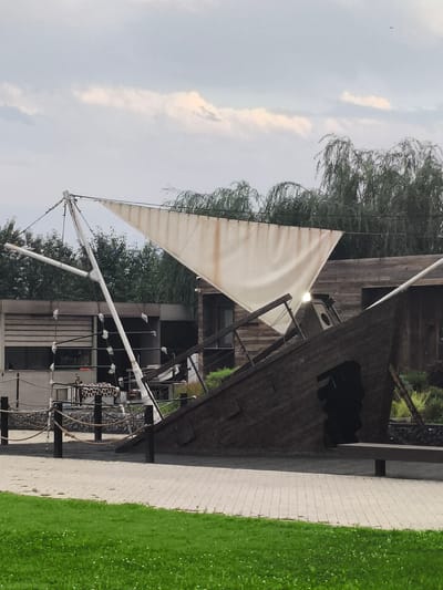 The image depicts a daytime scene in Komsomolskoe, Kyrgyzstan, featuring a unique, partially constructed ship-like structure with a large, white sail. The structure, made of dark wood, appears to be an artistic installation or a playground feature, tilted at an angle with an opening resembling a cave or entrance. A white mast supports the sail, which is stretched taut but shows signs of wear.

Behind the ship structure, there is a modern building with large windows and a roller-blinded section. A fence made of rope and posts separates the paved area from the grassy foreground. In the background, tall, weeping willow trees add a touch of nature to the scene. The sky is partly cloudy, suggesting fair weather.

The overall impression is one of a recreational area, possibly a park or an outdoor entertainment venue, where the ship-like structure serves as a focal point. There is no visible text in the image. The scene is quiet and still, with no people or active events depicted.