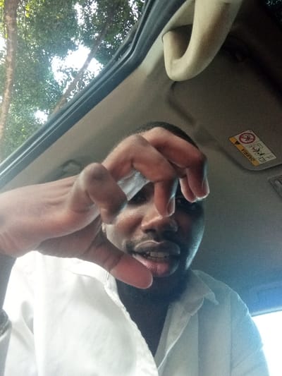 The image displays an adult male captured in a low-angle selfie from inside a vehicle. The individual, characterized by dark skin and short facial hair, is wearing a white collared shirt. He looks directly at the camera with a slight smirk. His right hand is prominently featured in the foreground, forming a 'pinching' or 'claw' gesture with partially curled fingers. The vehicle's interior ceiling is visible above and behind the subject, consisting of a light-colored headliner and dark trim. A beige overhead grab handle and a section of a sun visor, displaying an inverted red and yellow airbag warning label, are visible on the upper right. Through an apparent overhead opening, likely a sunroof, green tree foliage and branches are visible against a bright sky. The specific location within Makadara/Mji Wa Kale ward, Kenya, cannot be determined from the visual information present in the image.