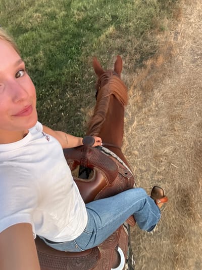 A high-angle perspective captures a female individual mounted on a chestnut-colored horse in an outdoor setting within Rancho Murieta, United States. The female subject, positioned in the left foreground, has light-colored hair and a fair complexion, and is looking towards the camera with a slight smile. She is wearing a white t-shirt, blue jeans, and light-colored, heeled boots. Her left hand rests on the pommel of a brown, intricately tooled Western saddle. A small linear tattoo is visible on her left forearm. The horse faces away from the viewer, its head and upper back visible, with a neatly laid mane. The ground surface displays a clear delineation between green, overgrown grass on the left and dry, straw-colored grass on the right, consistent with an open field or trail. Natural daylight illuminates the scene.