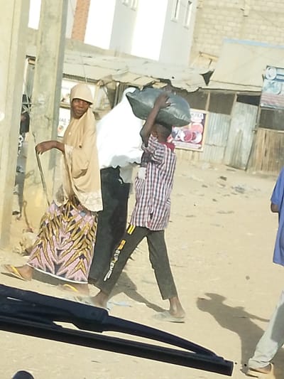 On a dusty street, a woman in a tan head covering and patterned skirt walks with a cane. Beside her, a child carries a large black bag atop their head. The child wears a plaid shirt and dark pants with white lettering. Buildings and shops line the background, and a glimpse of a car windshield is visible in the foreground. Another person in a blue shirt walks on the right side of the image.