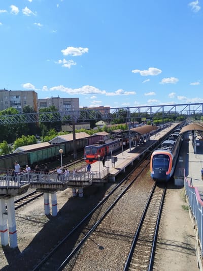 The image depicts a railway station scene in Monino, Russia, under a bright blue sky with scattered white clouds. The time of day appears to be mid-morning or early afternoon, judging by the strong sunlight casting distinct shadows.

The foreground features a pedestrian overpass with people walking across it. The overpass is supported by concrete pillars with red and grey markings. Below the overpass are multiple railway tracks.

Two trains are visible on the tracks. One is a red and grey passenger train stopped at a platform. Another is a modern, colorful passenger train, primarily blue and orange, with the Russian Railways logo on its front, positioned on a different track and appearing to be in motion or recently arrived. Several platforms with arched roofs are present, providing shelter for waiting passengers.

In the background, residential buildings and trees can be seen, indicating the station is situated in an urban or suburban area. The overall atmosphere is one of daily activity at a functioning train station. There is some visible text on the colorful train which appears to be Cyrillic, but it is too small to read clearly.