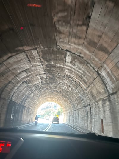 The media file captures a view from inside a wooden-lined tunnel. The tunnel walls are constructed from weathered wooden planks, showing signs of age and wear with visible cracks and discoloration. The perspective is looking forward towards the tunnel's exit, where daylight streams in, illuminating the road ahead.
The scene shows traffic within the tunnel. A car is visible further ahead, followed by at least one motorcycle. The road surface appears to be asphalt with white lane markings. Outside the tunnel's exit, trees and foliage are visible, suggesting a natural, possibly mountainous, environment. The lighting conditions indicate it is daytime, with the bright light at the exit contrasting with the dimmer interior of the tunnel.
There is a hint of a digital display on the right side of the frame, showing some red illuminated text, but it is largely unreadable due to the angle and resolution. The image is a still photograph, likely taken from a moving vehicle, as indicated by the slight motion blur in some areas of the wooden tunnel walls. The overall impression is of transit through a rustic and possibly historic tunnel. The location context is Prijevorac, Montenegro.