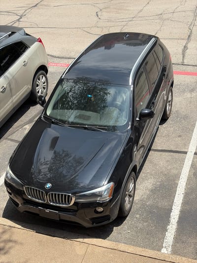 A black BMW X3 is parked in a paved parking lot in Grapevine, Texas. The vehicle is oriented diagonally, facing towards the lower left of the frame. A portion of a light-colored SUV is visible to the left of the BMW, and a red line is painted on the pavement between the two vehicles, indicating a parking space boundary. The parking lot surface shows cracks and wear. The BMW has its headlights on and a license plate is attached to the front bumper. Trees are reflected in the windshield of the BMW.