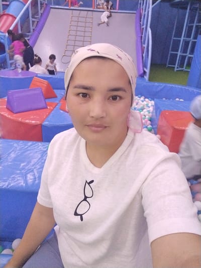 A medium shot photograph shows a woman wearing a white t-shirt with a glasses graphic on it and a white headscarf. She is in what appears to be a children's indoor play area in Talas City, Kyrgyzstan. The background is blurred but shows colorful soft play structures, a slide, and a ball pit. There are other people, likely children, in the background, some playing. The overall scene suggests a public recreation space for families.