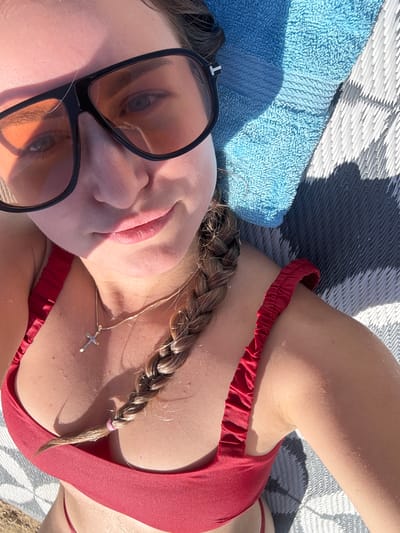 A young woman wearing a red bikini and large sunglasses lays on a striped mat outdoors. A blue towel is placed behind her head. Her hair is braided and falls over her shoulder. She is wearing a silver cross necklace. The lighting suggests it is daytime and sunny.