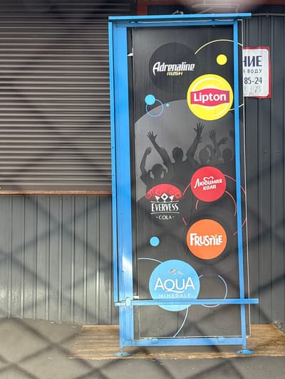 Here's a description of the image:
The image depicts an advertisement sign in Ust-Ordynsky, Russia. The sign has a blue frame. The background is dark, with silhouetted figures. There are brand logos, including "Adrenaline Rush," "Lipton," "EVERVESS - COLA," "Любимая кола" (Beloved Cola), "AQUA MINERALE," and "FRUSTILE." Above the sign, there is a partial sign in Russian, possibly indicating "НИЕ," "Я ВОДУ" and "85-24". The sign is mounted on a wall with a dark metal roll-up door and a gray horizontal panel wall. The adjacent ground area shows traces of moisture.