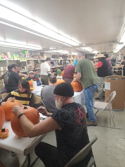 The image captures an indoor pumpkin carving event taking place in the city of Fife, United States. Approximately a dozen individuals, predominantly adults, are engaged in preparing and carving large orange pumpkins spread across white rectangular folding tables. The setting appears to be a large workshop or storage area, characterized by extensive metal shelving units stocked with various boxed and packaged items visible in the background. Overhead fluorescent tube lighting illuminates the scene.

In the foreground, a man with a white beard and a black beanie, wearing a black t-shirt with a red design on the back, is seated at a table, actively marking a pumpkin with a small tool. To his left, a person wearing a yellow and black striped top is seated, observing a pumpkin. Further into the scene, individuals in various attire, including a man in a green t-shirt and blue jeans, and another wearing a conical straw hat, are focused on their respective pumpkins. A large, light brown cylindrical barrel stands on the right side of the image. The flooring is a light-colored, industrial-style surface. People are primarily focused on the task of carving, with no specific inter-personal interactions highlighted.