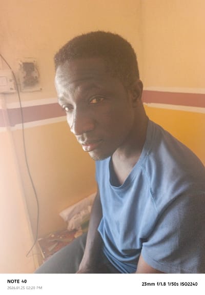 A male individual is depicted indoors in Tudun Wazurchi, Nigeria, on January 25, 2026, at 02:20 PM. The subject, an adult with dark skin and short dark hair, is positioned slightly off-center to the right, looking directly towards the camera with a neutral expression. He wears a blue, short-sleeved, crew-neck t-shirt and dark trousers. A linear facial scar is visible on his left cheek. The background consists of a cream-colored wall painted with a horizontal reddish-brown stripe at mid-height. On the left side of the frame, a white light switch panel with exposed wiring is affixed to the wall, below which a discolored or damaged patch is evident. A diffused light source, originating from the left, illuminates the scene, casting a warm glow and creating a lens flare effect on the left. Indistinct objects, including a crumpled white cloth, are visible on a surface in the lower-left foreground. The image was captured with a NOTE 40 device using 23mm f/1.8 optics at 1/50s shutter speed and ISO 2240.