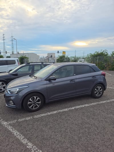 A grey Hyundai i20 is parked in an outdoor parking lot. The car is facing right, and its front is slightly angled towards the camera. The car is wet, suggesting recent rain. In the background, two other cars are visible: a dark-colored station wagon and a white van. Beyond the parking lot, there are power lines, industrial buildings, and trees. The sky is overcast with a bright patch of sunlight. The location is identified as Worms, Germany.