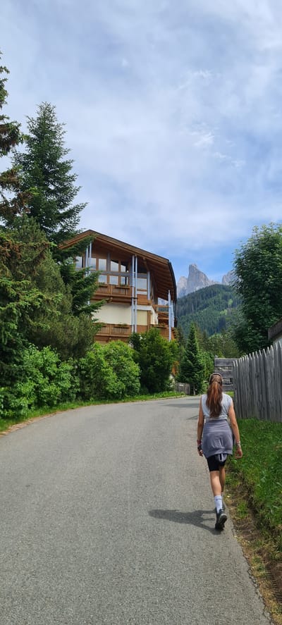 In St. Magdalena, South Tyrol, Italy, a woman walks away from the camera on a paved road. She has long brown hair, wears a grey t-shirt, black shorts, and white socks with black shoes, and has a grey jacket tied around her waist. The road is lined with green grass and a wooden fence on the right side. On the left, a multi-story wooden and glass building is visible, surrounded by lush green trees. In the background, a dramatic mountain peak rises against a partly cloudy sky.