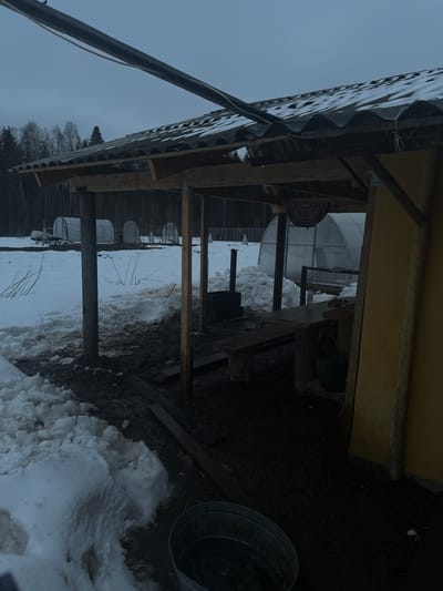 The image depicts an outdoor scene in Zapadnaya, Russia, under an overcast sky. The ground is partially covered in snow, with visible patches of mud. The primary structure is a wooden shed or shelter with a corrugated metal roof. Supporting the roof are wooden beams and metal posts. Behind the shed, several greenhouse structures are visible. A metal bucket is present near the edge of the shed. The lighting is dim, and the overall scene appears rural or semi-rural. There is no visible presence of people.