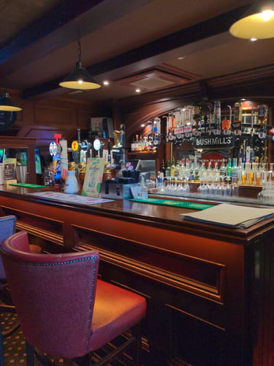 This is an indoor shot of a bar, likely taken in the evening given the lighting. The scene is dominated by a dark wooden bar, laden with bottles of alcohol and beer taps. A prominent "BUSHMILLS IRISH WHISKY" sign is visible behind the bar, along with many other brands. Several beer taps with their associated labels are also visible, including "SPELL", "GUINNESS", and "OUTCIDER". A red upholstered bar stool is in the foreground, facing the bar. The overall atmosphere appears to be that of a traditional pub or bar, with warm lighting and rich wood tones. There are no people visible in the image.