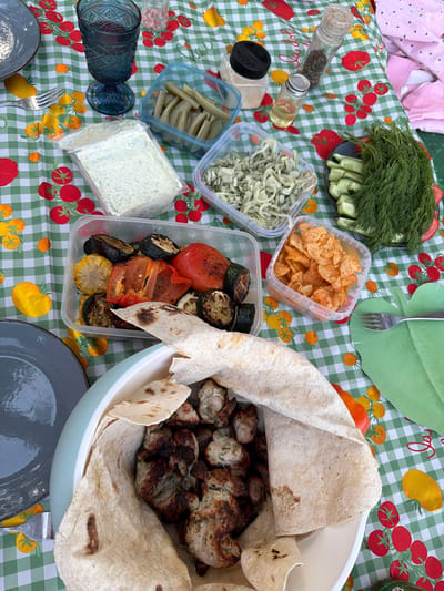 This image depicts a picnic table laden with food. The table is covered with a checkered tablecloth patterned with tomatoes and yellow vegetables. Various food items are arranged on the table, including a bowl filled with grilled meat and flatbread, plastic containers holding pickles, cabbage salad, grilled vegetables, chips, dill and cucumbers, and a block of cheese. There are also bottles of spices. Plates and forks are set on the table. The location is likely Plieņciems, Latvia, given the presence of food items and setting typical of a picnic. There are no people visible in the scene.