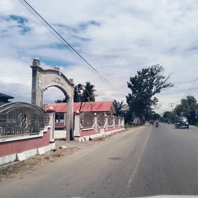 This daytime image captures a road scene in Keudealuerheung, Indonesia, under a sky with scattered clouds. The photograph appears to be taken from inside a vehicle, looking forward.

On the left side of the road, a substantial concrete archway serves as an entrance, featuring decorative elements like small dome-shaped finials and embedded lights. Inscribed on the archway is the text "MEUNSAH KEUDE ALUEREUNG". Adjacent to the archway, a decorative fence with ornate metalwork atop a red-painted lower wall extends along the roadside. Behind this fence and archway, a building with a distinctive red corrugated metal roof is visible, partially obscured by palm trees.

The asphalt road, which appears dry, stretches into the distance, showing faint white road markings in the foreground. Traffic is present on the road; a dark-colored pickup truck or van is driving towards the viewer on the right side of the road, while several motorcycles or scooters are further down the road, traveling away from the camera.

The environment is characterized by lush greenery, with numerous trees lining both sides of the road, including the prominent palm trees near the building and a large, leafy tree with some dead branches on the right. Utility poles and electrical wires are visible across the top of the frame and along the roadside, indicating developed infrastructure. The lighting suggests it is late morning or early afternoon, with clear visibility. The scene depicts an ordinary moment of daily life and transit in the area.