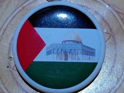 A round pendant displays colors mimicking the Palestinian flag: black, white, red, and green. The white stripe features an image of the Dome of the Rock in Jerusalem. The red is a triangle shape. The pendant has a white plastic rim and a metal loop at the top. The pendant rests on a textured, light-colored surface.