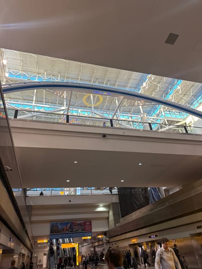 The image captures the interior of an airport terminal, showcasing a curved glass wall with structural supports and a circular yellow element. Adjacent to the glass structure is a broad, light-colored wall with small, evenly spaced lights. The scene extends into a bustling area with escalators, passengers, and signage. An overhead structure with linear elements creates a sense of depth and perspective.