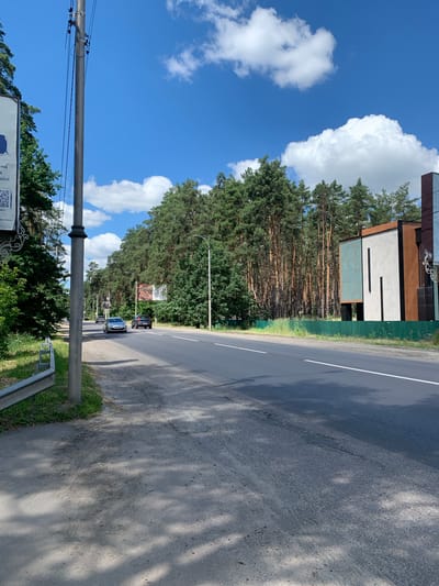 This image depicts a street scene in Bucha, Ukraine, on a sunny day with scattered clouds. The perspective is from the side of a paved road, looking forward.
In the foreground, on the left, is a paved sidewalk adjacent to a metal guardrail. A concrete utility pole with wires runs vertically along the left side of the frame. Further down the road on the left, a white sign with text and a QR code is partially visible.
The road itself is asphalt with a white dividing line. Two cars are traveling on the road; a silver sedan in the left lane and a dark SUV in the right lane.
The environment is characterized by a dense line of tall pine trees forming a backdrop along the right side of the road. Behind the trees, on the far right, a modern building with a geometric design, featuring panels of white, brown, and teal, is visible. A green fence runs along the edge of the property where the building is located. The sky is blue with several fluffy white clouds. The scene appears tranquil and ordinary, showing everyday life on a street in Bucha.