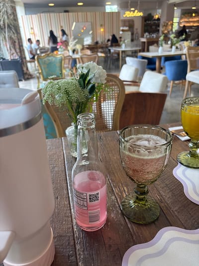 The image shows a close-up of a table in what appears to be a restaurant or cafe in Rionegro, Colombia. In the foreground, there is a light pink appliance, a glass bottle with pink liquid and a label with nutritional information and a QR code, and a green glass filled with a pink bubbly beverage. To the right of that, another green glass holds a yellow liquid. A bouquet of white flowers and greenery is in a small glass vase behind the bottle. The table is made of dark wood planks. In the background, the restaurant's interior is visible, with striped wallpaper, chairs, tables, and patrons. A neon sign that says "Feliciter" is mounted on the wall.