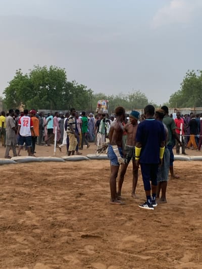 The image shows a group of people gathered around a wrestling ring in Sokoto, Nigeria. The ring is located at 13.049 latitude and 5.238 longitude. The ring is made of dirt and surrounded by a ring of sandbags. There are numerous people gathered around the ring. Five men stand in the center of the ring. They are all shirtless and wearing shorts. One of the men is wearing a blue and white striped shirt, another is wearing a black and red shirt, and another is wearing a yellow shirt. The men appear to be involved in a wrestling match. There are many spectators watching the wrestling match. It is daytime and the sky is cloudy.